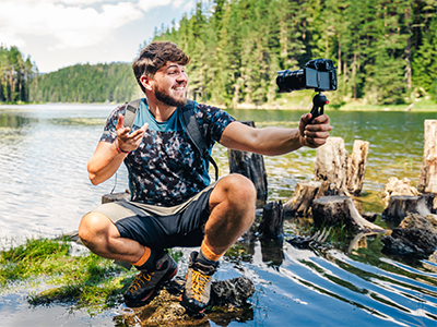 Male student poses with a video camera while perched on a log close to shore on a lake.