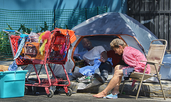 Middle age Black man lounging in a tent talking to a older White woman sitting in a lawn chair just off a sidewalk