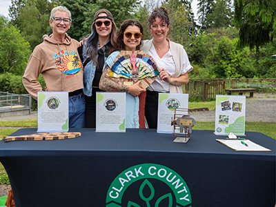 Four female Public Health employees stand behind a table with a banner featuring the Clark County Green Business logo. Samples of the Green Business awards are featured on the table.