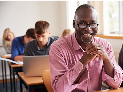 Middle-aged Black man sits at a desk in a classroom with other students. He is smiling and looking directly at the camera.