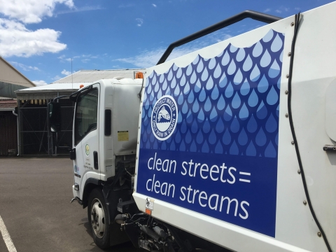 Employee wearing orange high visibility vest drives a white street sweeper with the Clark County logo seen on the driver side door.