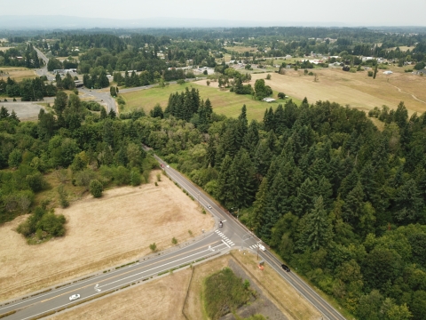 Aerial photo showing brown grass fields with stand of trees scattered among them and roads running through them. Some buildings are seen in the midground, and there are hills and a gray sky in the background.