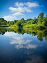 Lush, green deciduous trees, a blue sky and a few small clouds are reflected in the still water of the river in the foreground.