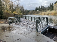 A platform over a rushing river, with mesh decking, metal railing, surrounded by tree canopy. A cloudy sky is in the background.