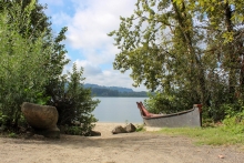 A tan colored, crushed gravel path is in the foreground, shrubs and deciduous trees and a traditional-style canoe are in the midground, and a sandy beach, river, hills, and partially cloudy sky are in the background. 