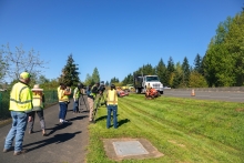 People in high-visibility reflective florescent colored vests observe a worker on a riding mower mowing a shoulder near a four lane high-speed roadway, on a cloudless day.