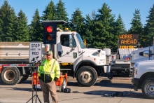 A worker wearing a reflective, florescent colored coat speaks at microphones with road construction trucks and an electronic message board reading "workers ahead" in the background, on a cloudless day.