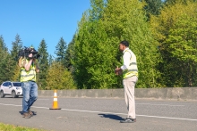 A man wearing a reflective florescent colored vest standing at the edge of the travel lane of a four-lane roadway speaks to a camera held by another person in a high-visibility vest on a cloudless spring day.