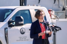 A woman in a pantsuit speaks at microphones set up in front of a white Clark County Public Works truck on a sunny day.